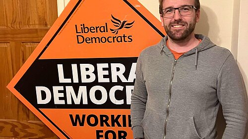 candidate, Pele Barnes, standing in front of a stakeboard saying Liberal Democrats, Working For You.